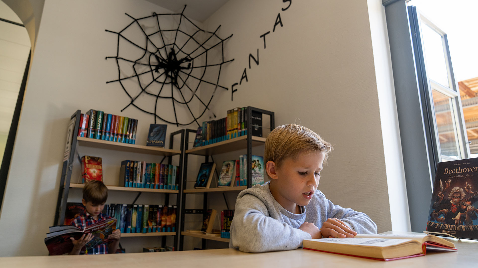 Ein kleinerer Junge liest in der Stadtbcherei in einem Buch, hinter ihm hngt ber einem Bcherregal ein groes schwarzes Spinnennetz mit dem Schriftzug Fantasy